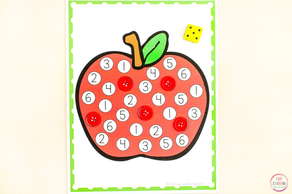 A large apple is on a sheet of paper with a green border. There are dots all over the apple and numbers on the dots. A dice is also placed on the mat.